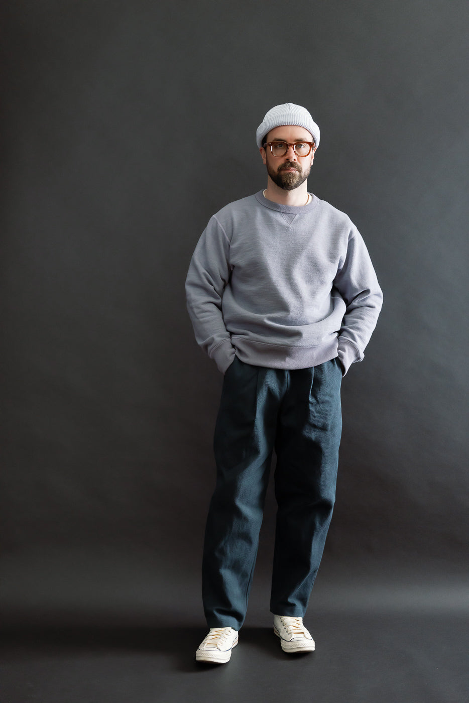 A man in a light gray sweatshirt and white beanie wears JM4623 - Sashiko Ump Pants - 280 Iron Navy, styled with pleated front and large pocket, showcasing their structured drape and raw denim-like appearance.