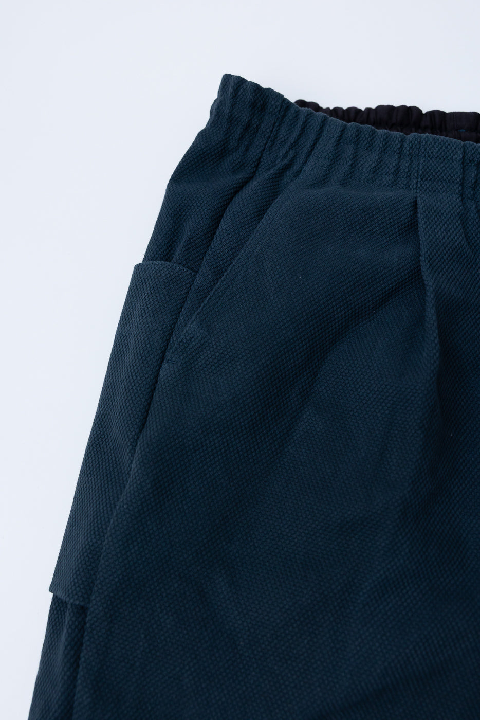 JM4623 - Sashiko Ump Pants - 280 Iron Navy: Close-up of dark navy pleated trousers with an elasticized waist, front pleat, and large side pocket, showcasing textured sashiko cotton fabric resembling raw denim.