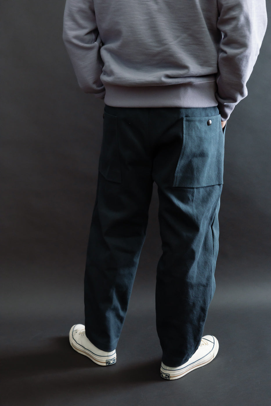 JM4623 - Sashiko Ump Pants - 280 Iron Navy, featuring large rear patch pockets with snap-button closures, worn with tucked light gray sweatshirt and white low-top canvas sneakers, showcasing a structured, raw denim-like drape.
