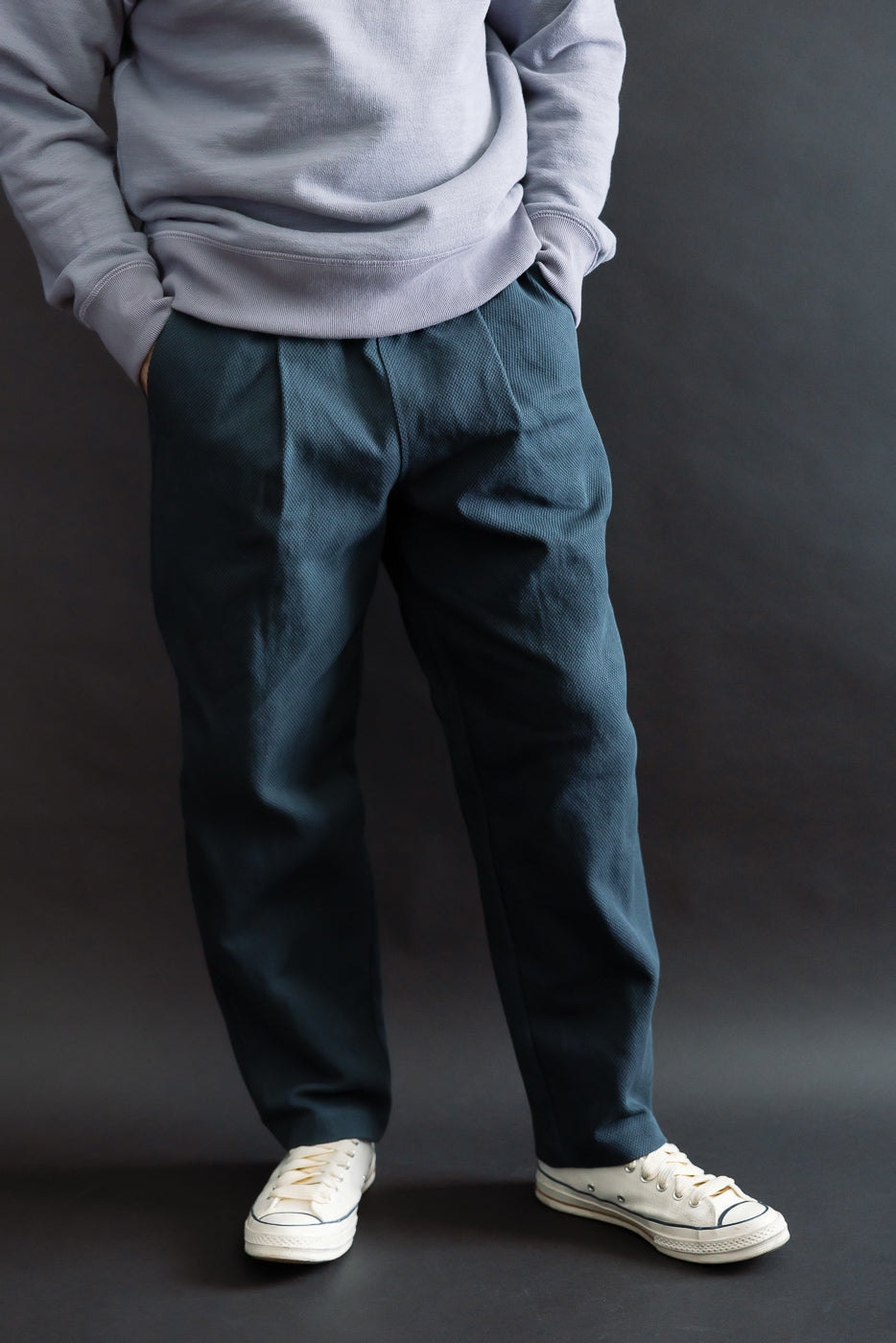JM4623 - Sashiko Ump Pants - 280 Iron Navy displayed on a model, showcasing relaxed-fit, pleated dark-blue trousers with large waist-hip pockets, paired with off-white canvas sneakers against a dark background.
