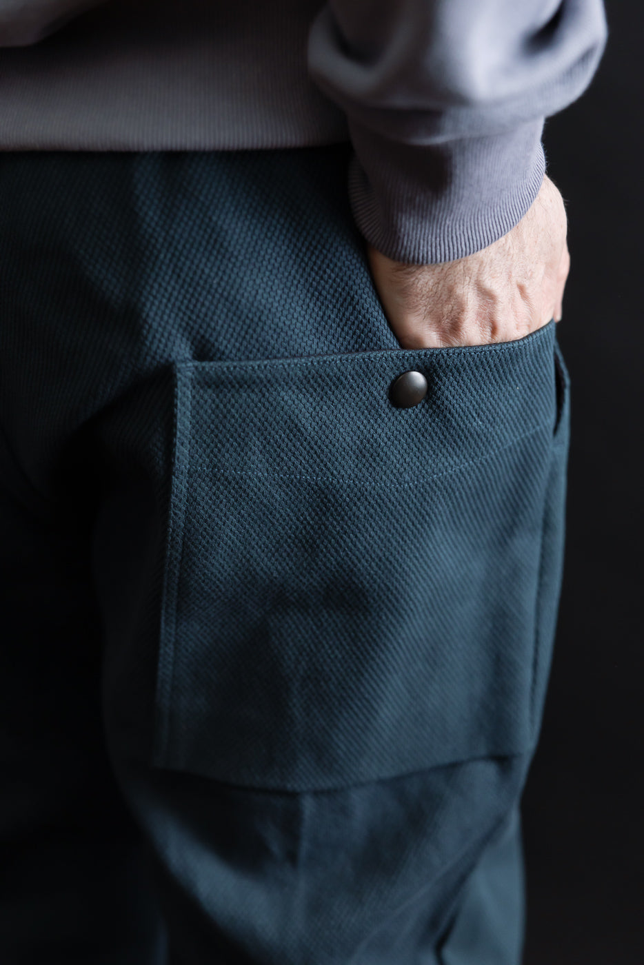 JM4623 - Sashiko Ump Pants - 280 Iron Navy close-up, showcasing textured teal sashiko fabric with a matte black snap button on the back pocket, worn with a grey top, emphasizing craftsmanship and structure.