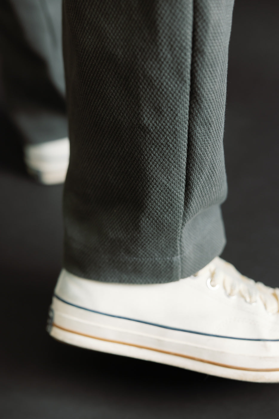 Close-up of JM4623 - Sashiko Ump Pants in dark textured fabric, showcasing detailed sashiko stitching and pleated front, paired with white canvas sneakers featuring a blue stripe along the sole.