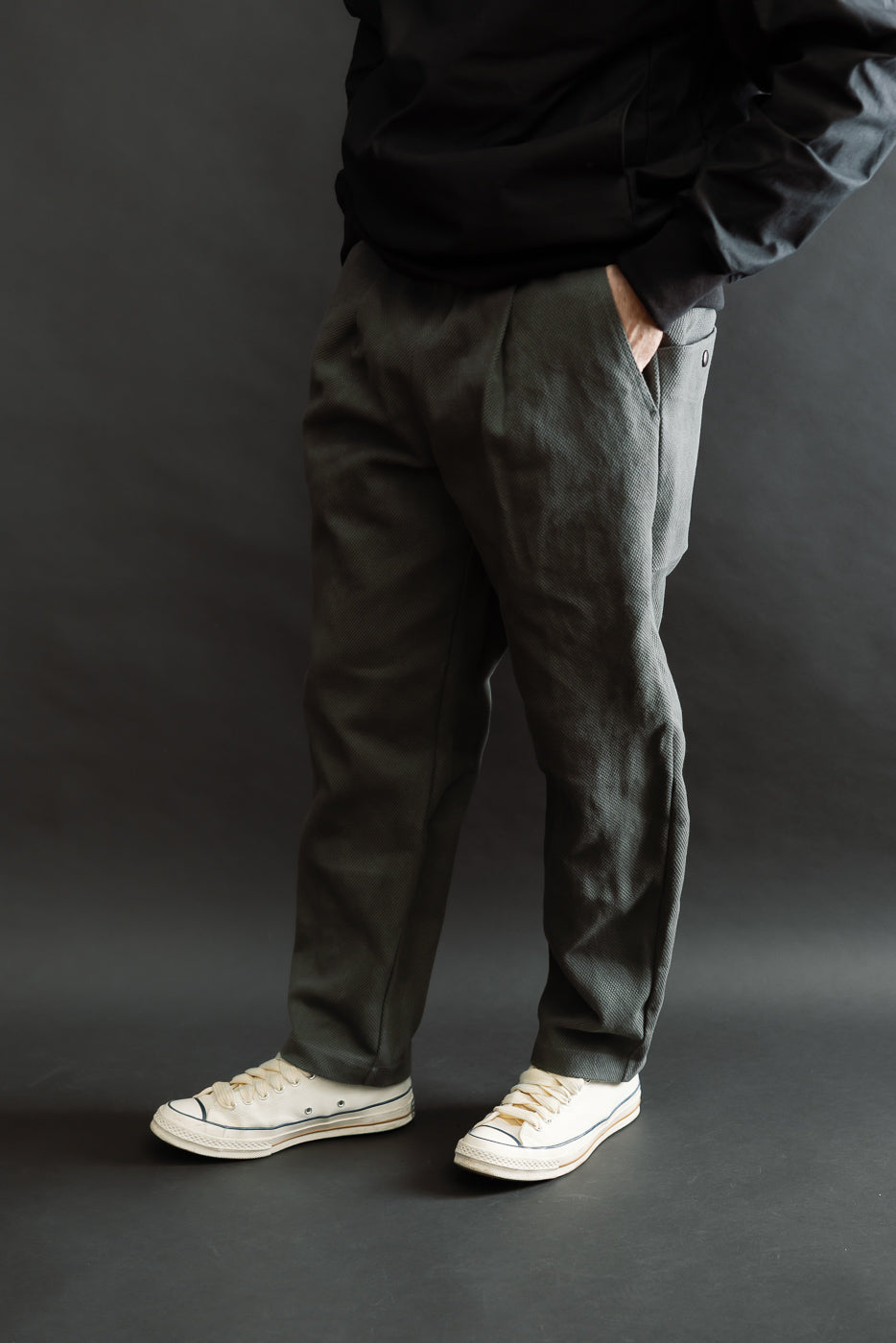 JM4623 - Sashiko Ump Pants - 333 Falcon Gray: Dark-gray textured trousers with a pleated front and large pocket, styled with off-white canvas sneakers against a dark backdrop, showcasing their structured drape and unique fabric.