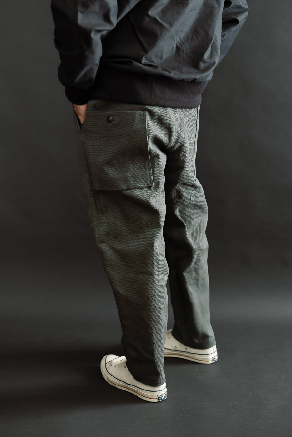 JM4623 - Sashiko Ump Pants in 333 Falcon Gray, featuring textured charcoal-gray fabric with a large patch pocket, viewed from the back, showcasing the unique drape and structure against a dark backdrop.