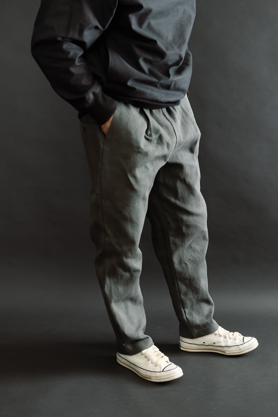 JM4623 - Sashiko Ump Pants - 333 Falcon Gray, featuring olive-gray textured pleated trousers with large pockets and exceptional drape, paired with off-white canvas sneakers for a refined, utilitarian look.