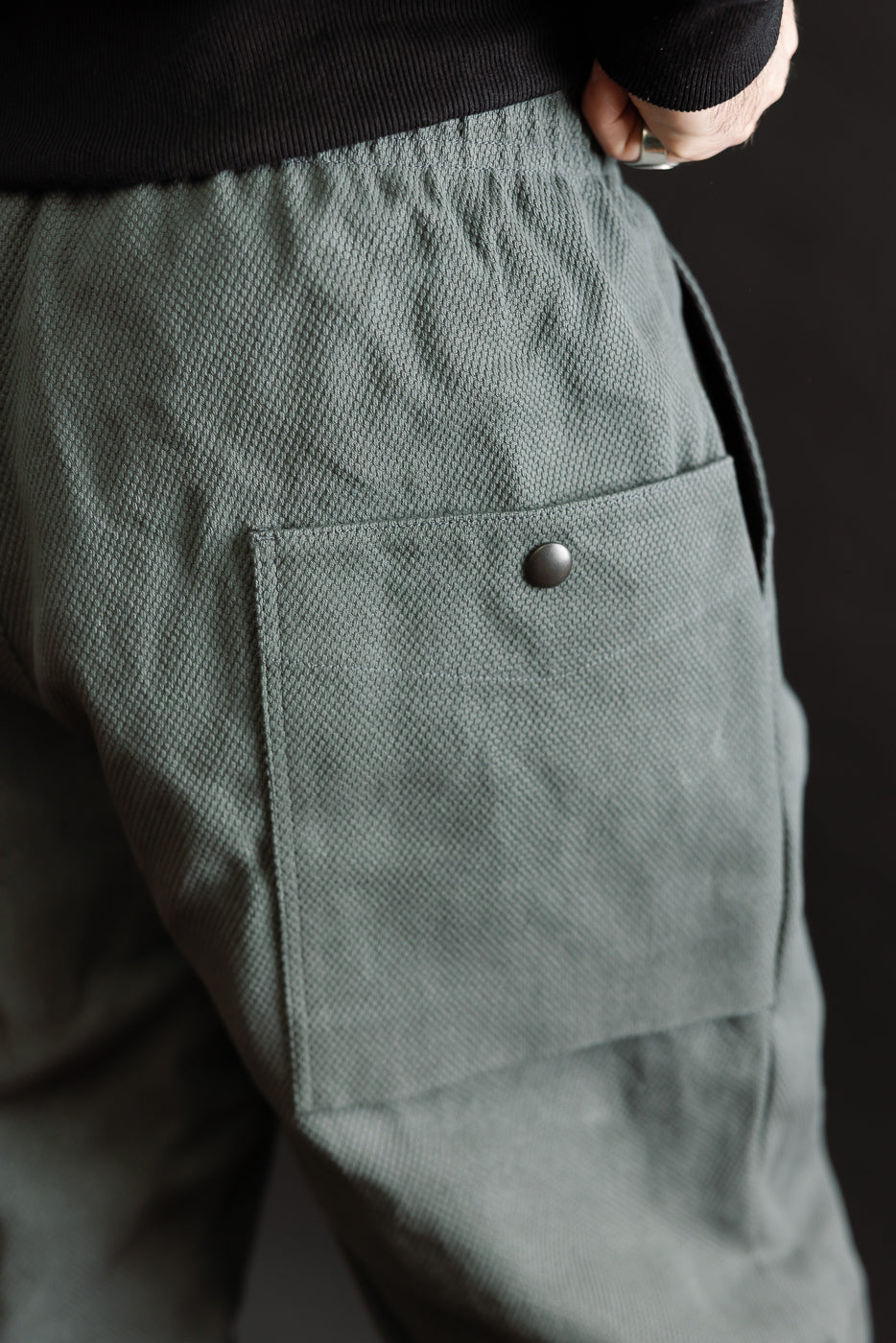 JM4623 - Sashiko Ump Pants in olive-green, showcasing textured sashiko fabric with an elasticated waistband and large flap pocket. The side-seam hand placement highlights the pants' utilitarian style and structured drape.