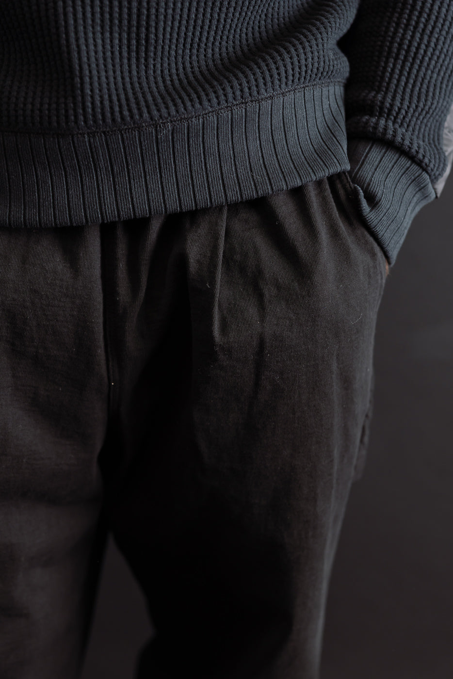 Close-up of the front side of a male model's black cotton trousers and ribbed knit sweater hem.