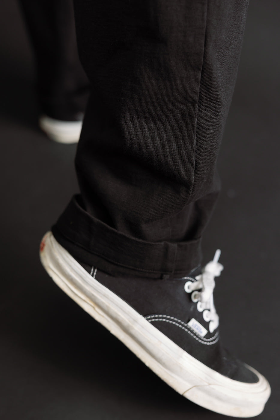Close-up of the cuffed hem of black trousers and black canvas sneakers on a male model.