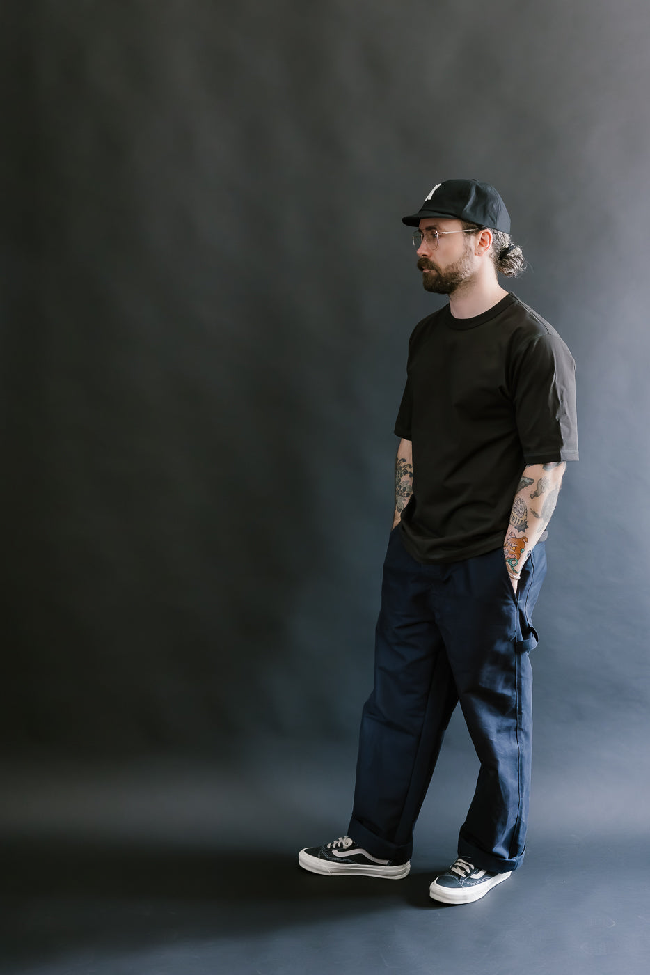 Man wearing a black t-shirt, dark pants, and a cap against a gray background