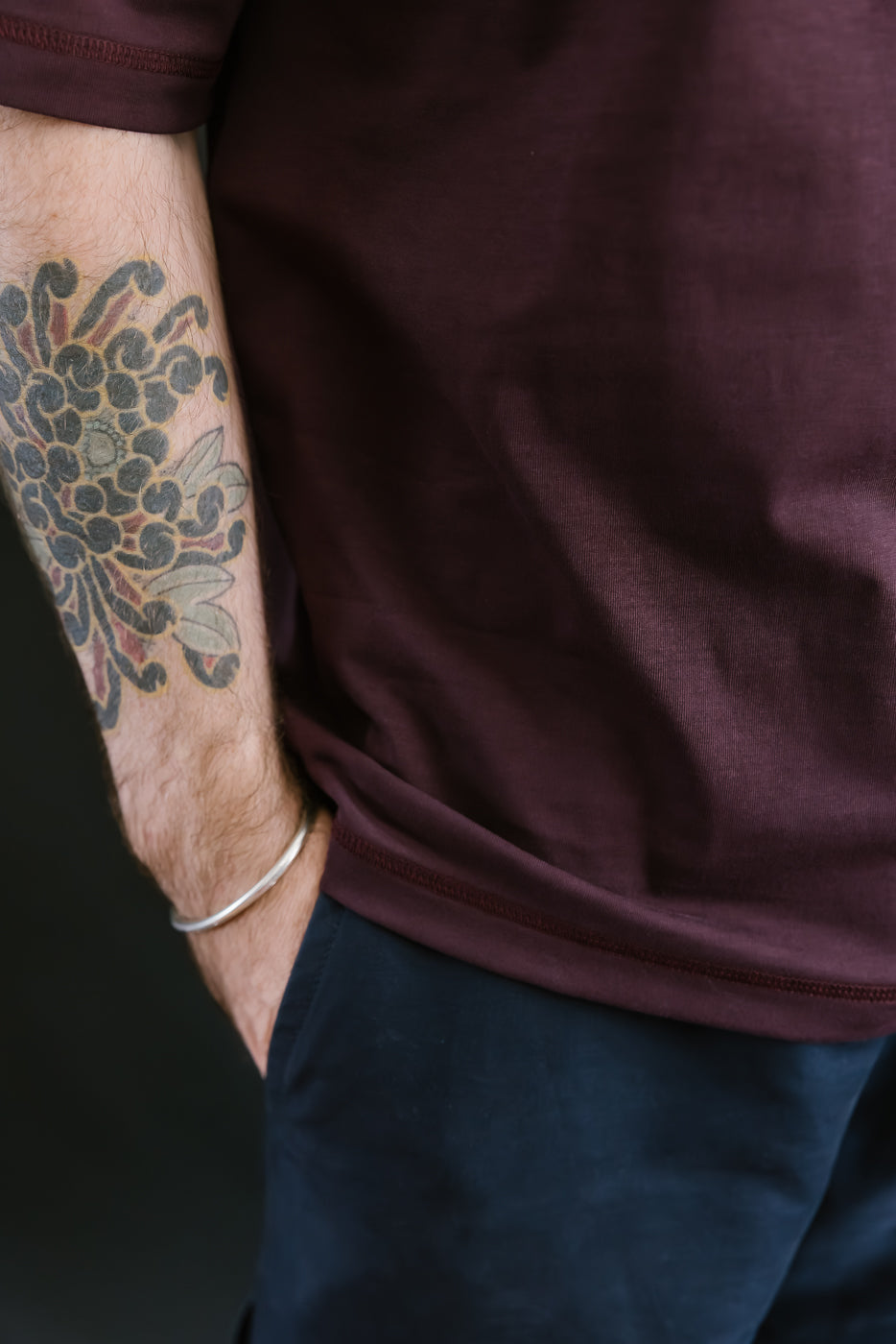 Person wearing a maroon shirt with a tattoo on their arm against a dark background