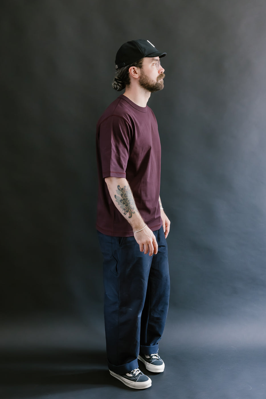 Man wearing a maroon t-shirt, dark pants, and a black cap against a gray background