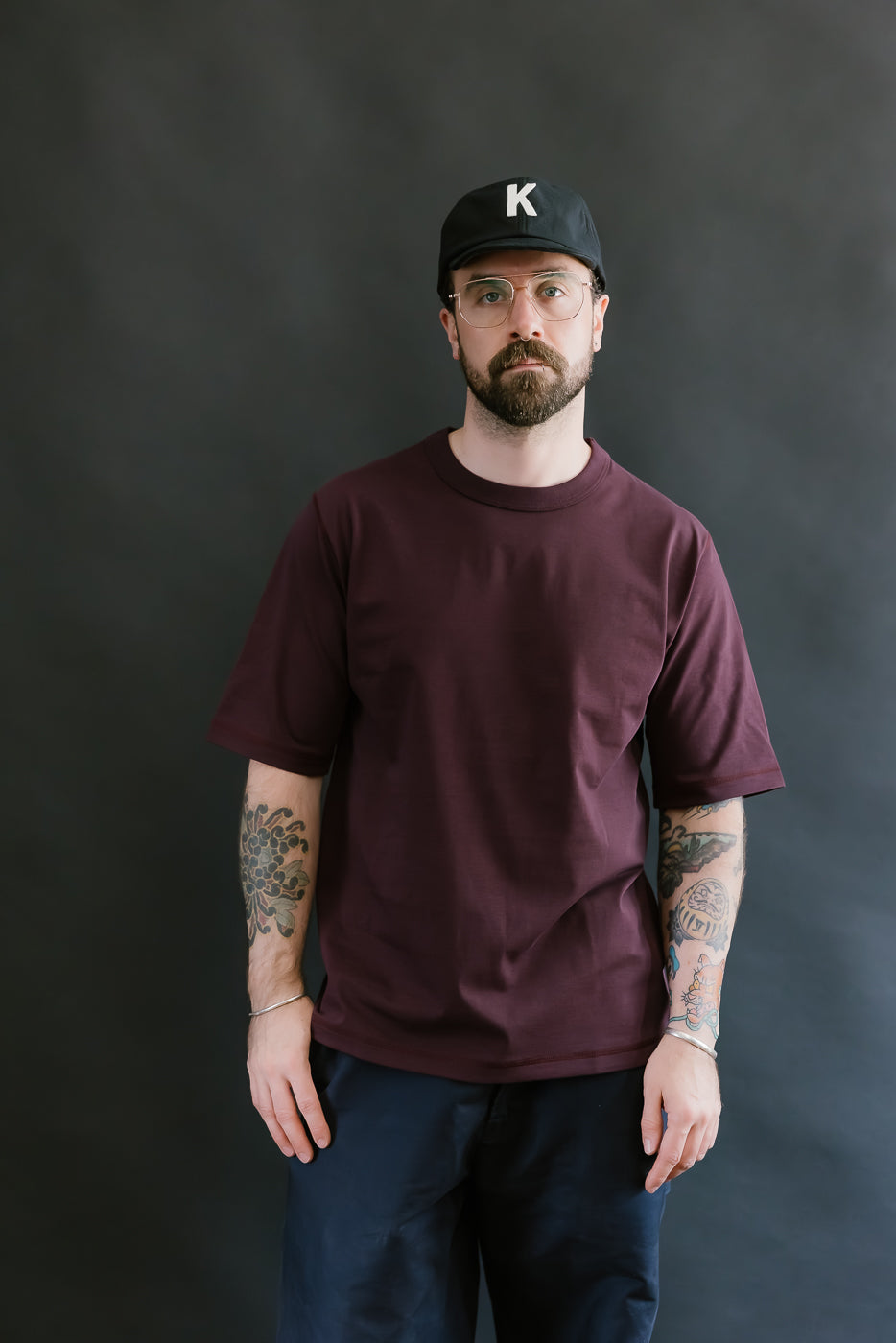 Man wearing a burgundy t-shirt and black cap with a 'K' logo against a dark background