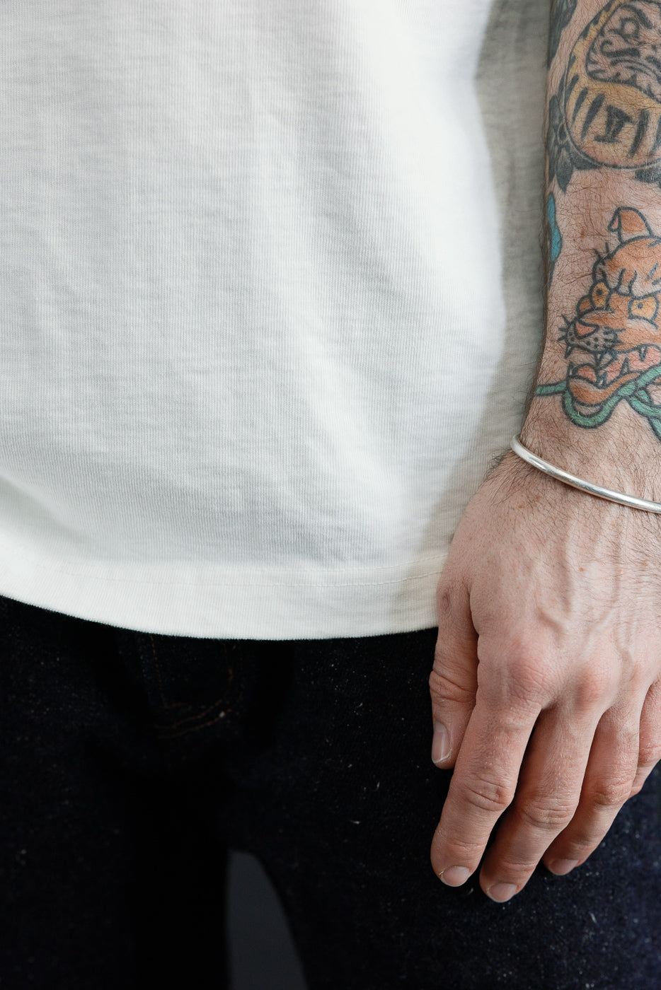 Close-up front view of male model’s torso and arm, showing white tee hem, tattooed forearm, and silver bracelet.