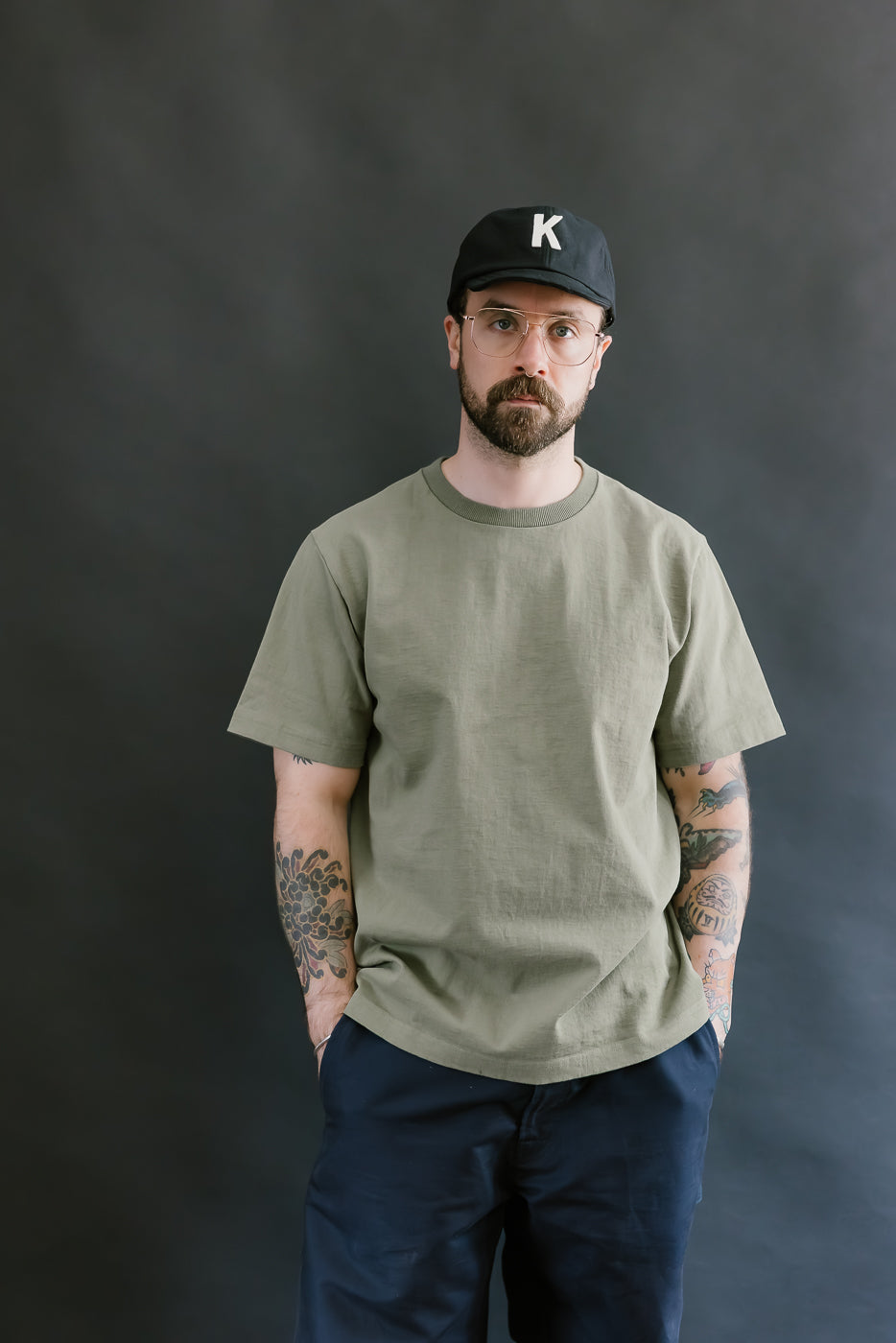 Man wearing a green t-shirt, navy pants, and a black cap with a 'K' logo against a dark background