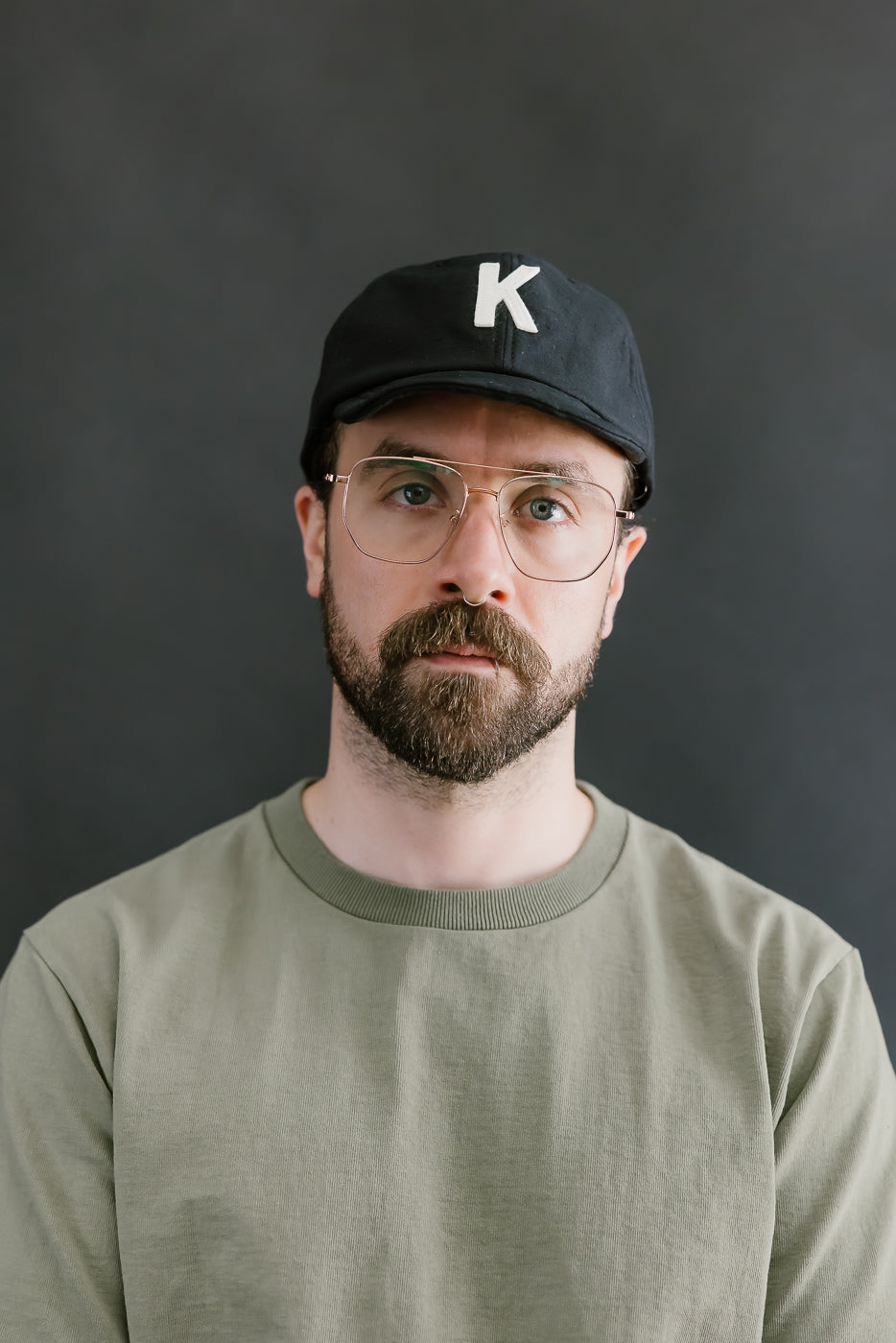 Man wearing a black cap with a white 'K' logo and glasses against a dark background