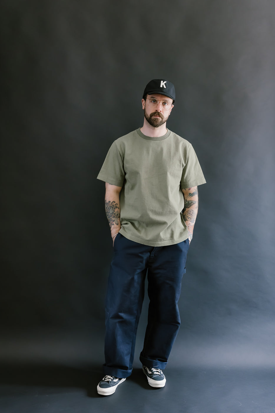 Man wearing a green t-shirt, blue pants, and a black cap with a logo against a dark background