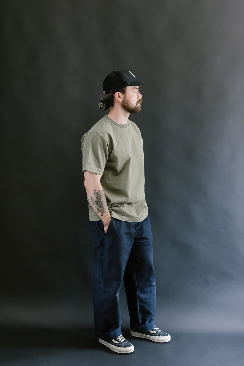 Man wearing a green t-shirt, blue jeans, and a black cap against a dark background
