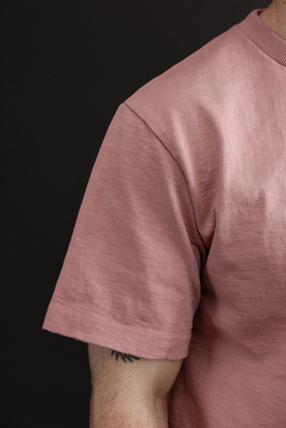 Close-up of male model's left shoulder and sleeve, showcasing pink t-shirt with ribbed texture and hem detail.