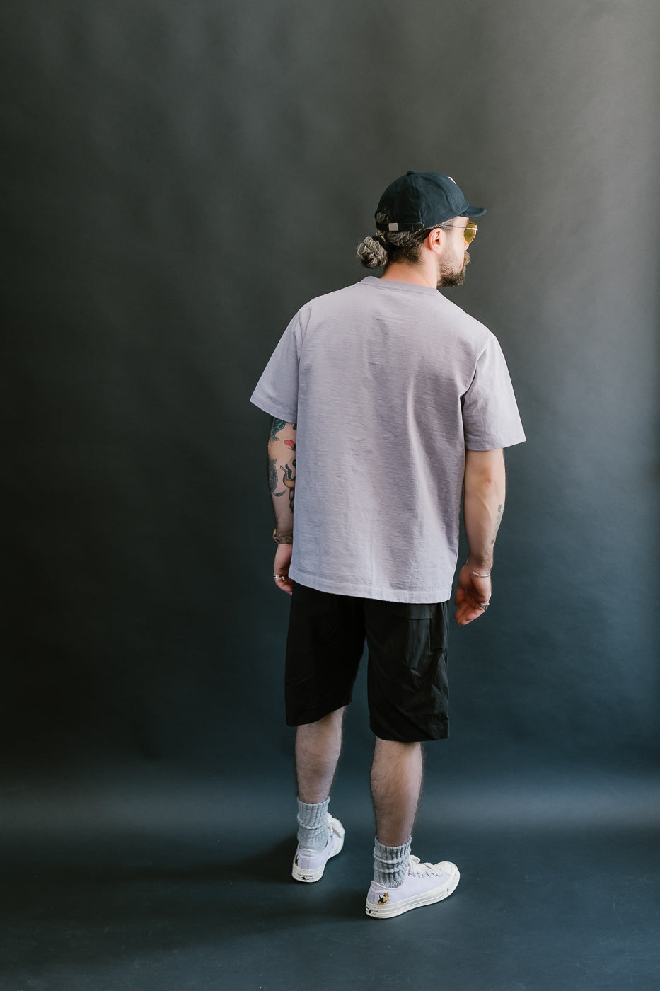 Back shot of person wearing a light purple t-shirt, black shorts, and light purple
 sneakers against a dark background