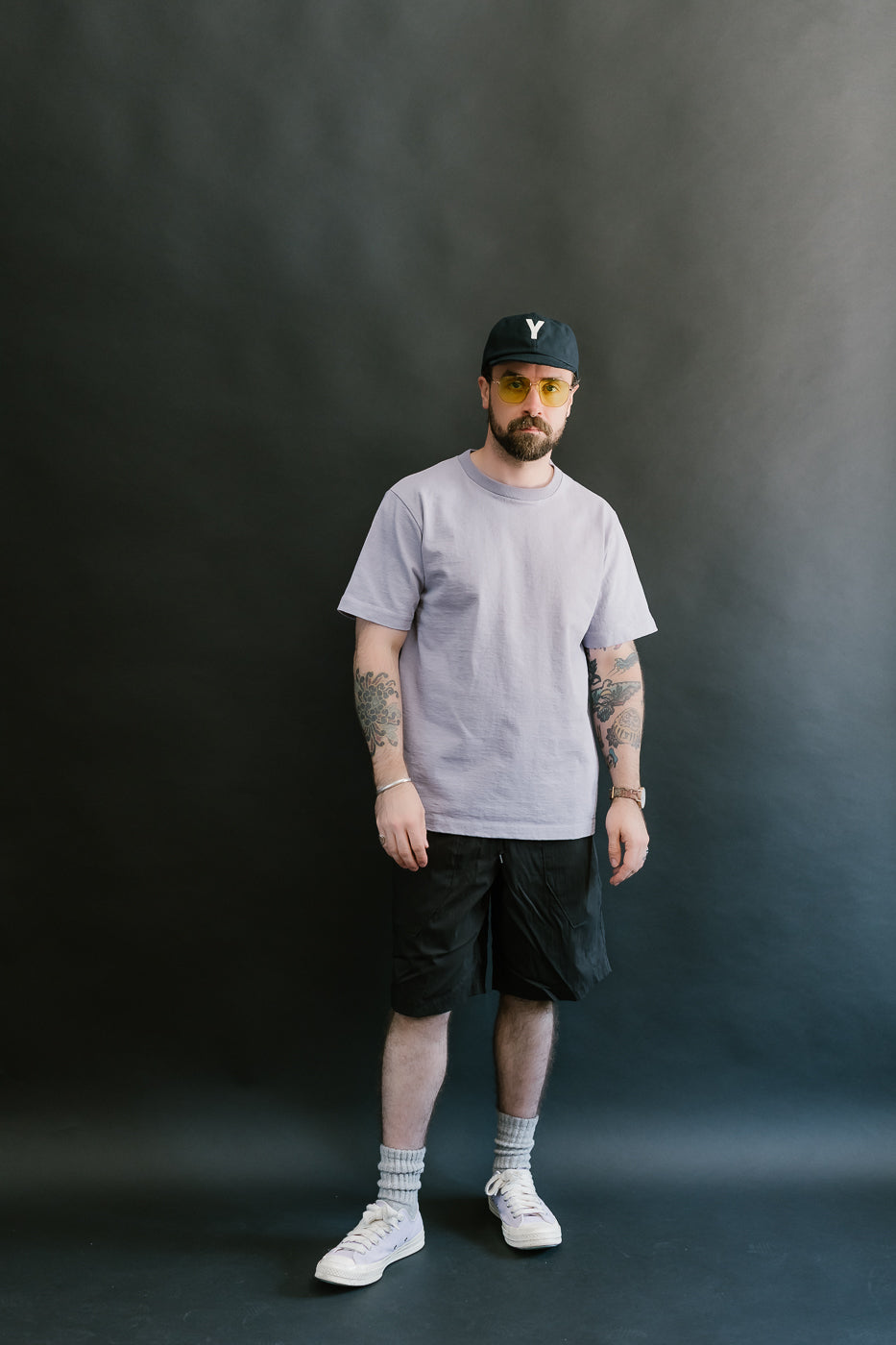 Man wearing a light purple t-shirt, black shorts, light purple sneakers, and a black cap against a dark background