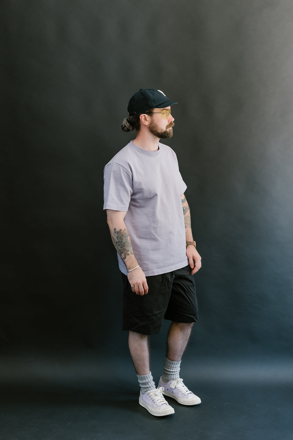 Man wearing a light purple t-shirt, black shorts, white sneakers, and a black cap against a dark background