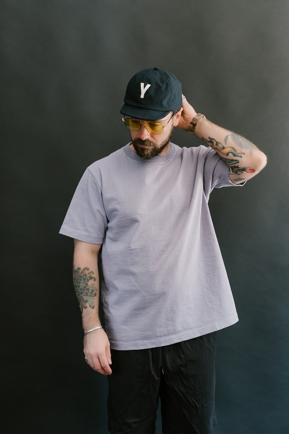 3/4 front view of a man wearing a light purple t-shirt, black cap, and sunglasses against a dark background