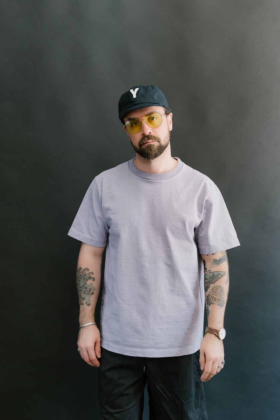 3/4 front view of a man wearing a light purple t-shirt, black cap, and sunglasses against a dark background