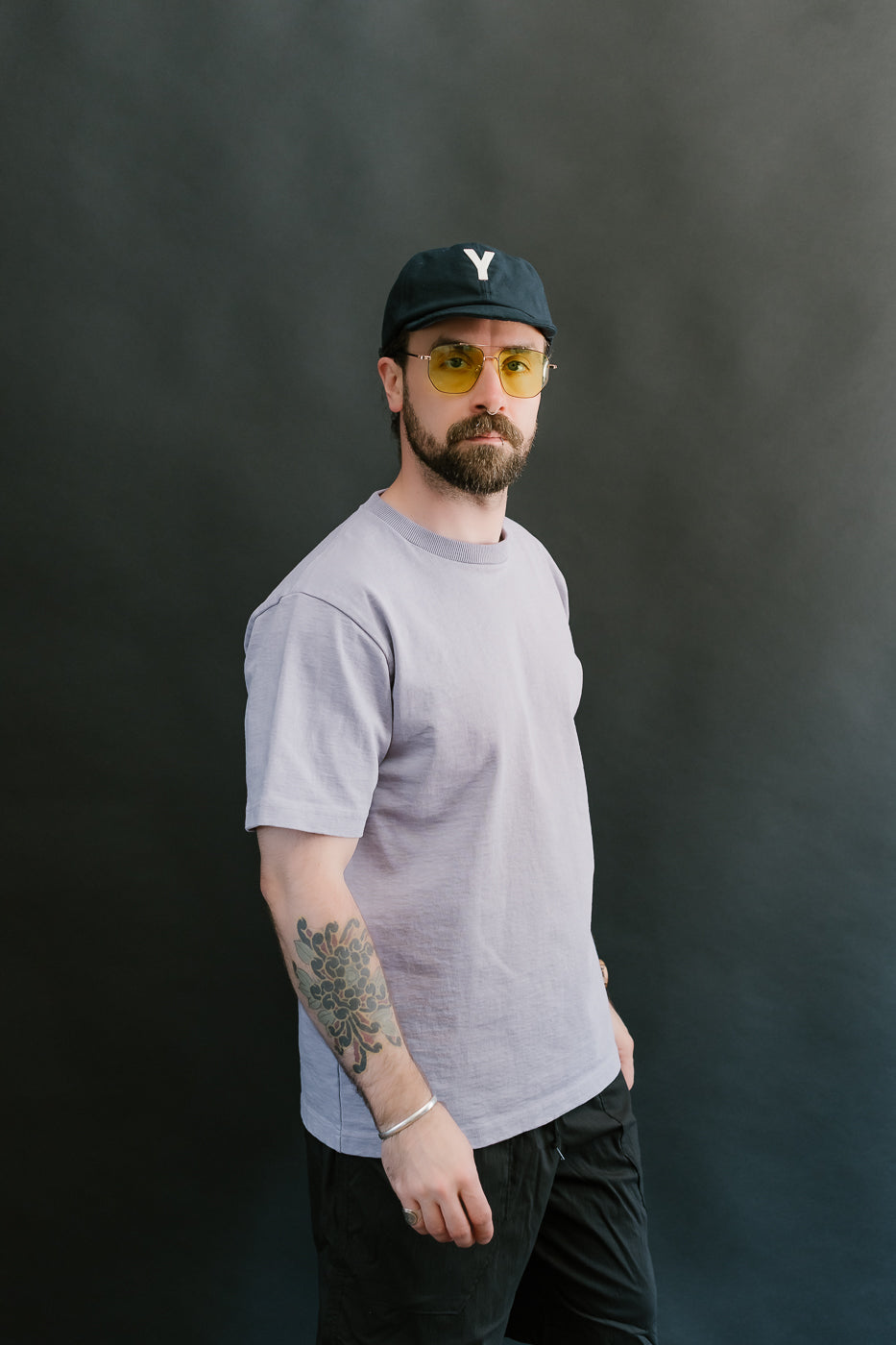 3/4 side view of a man wearing a light purple t-shirt, black cap, and sunglasses against a dark background