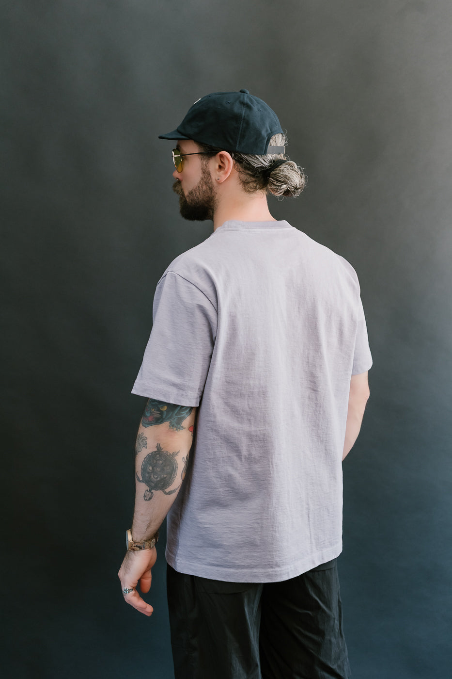 3/4 back view of a man wearing a light purple t-shirt, black cap, and sunglasses against a dark background