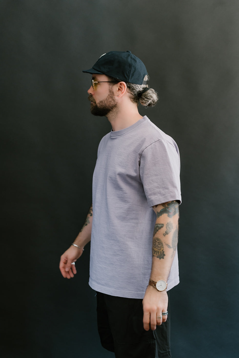 3/4 side view of a man wearing a light purple t-shirt, black cap, and sunglasses against a dark background