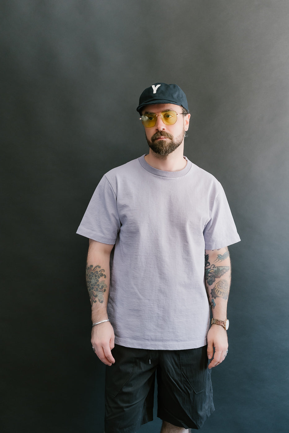 3/4 front view of a man wearing a light purple t-shirt, black cap, and sunglasses against a dark background