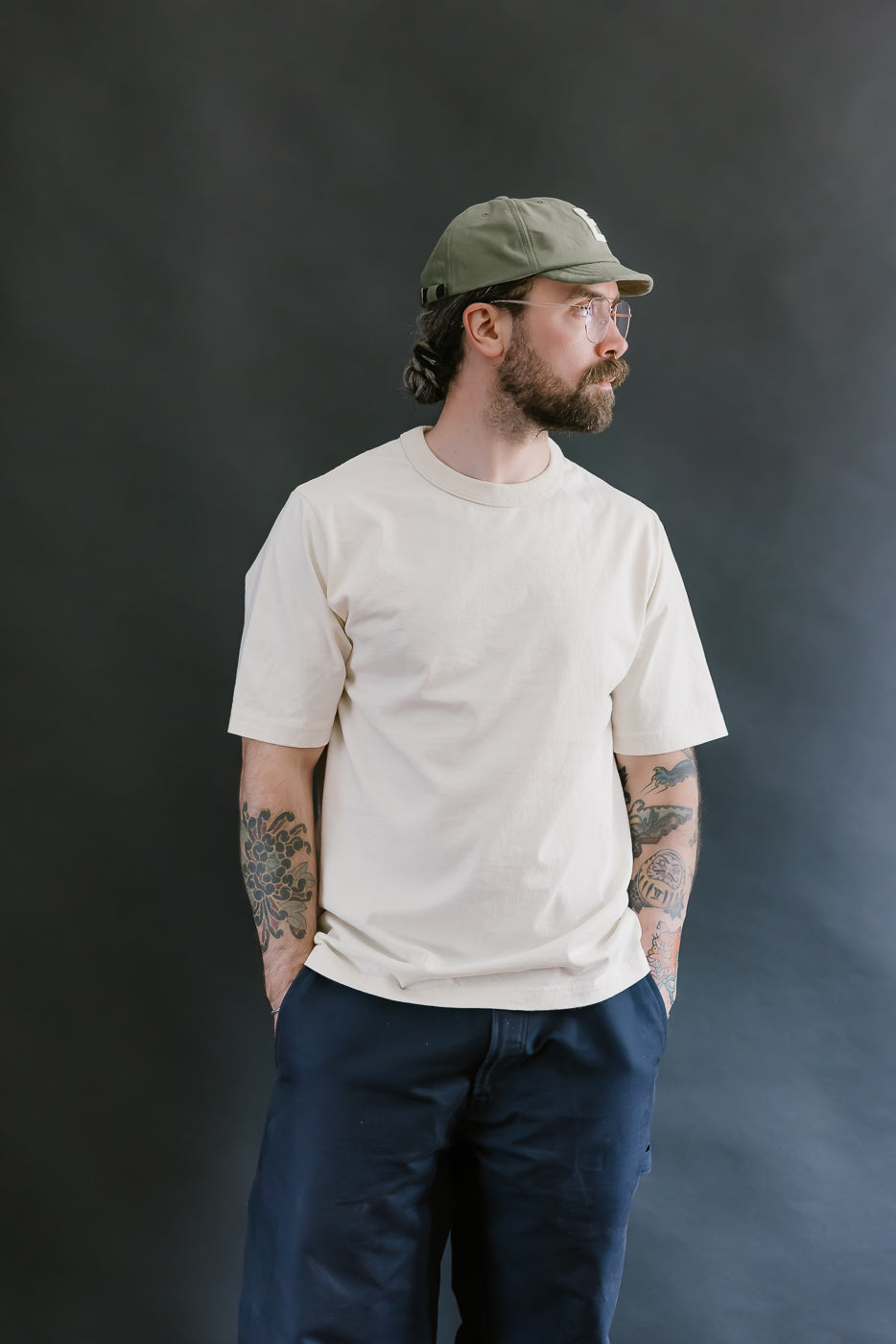 Man wearing a beige t-shirt, navy pants, and a green cap against a dark background