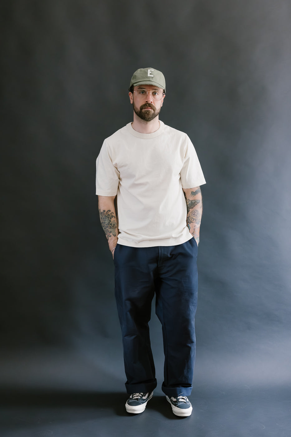 Man wearing a beige t-shirt, navy pants, and a cap against a dark background