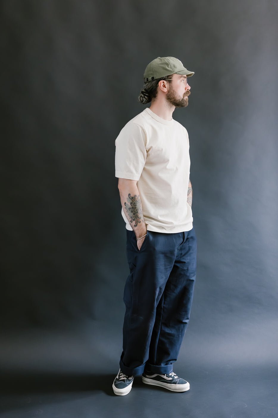 Man wearing a beige t-shirt, navy pants, and a cap against a dark background