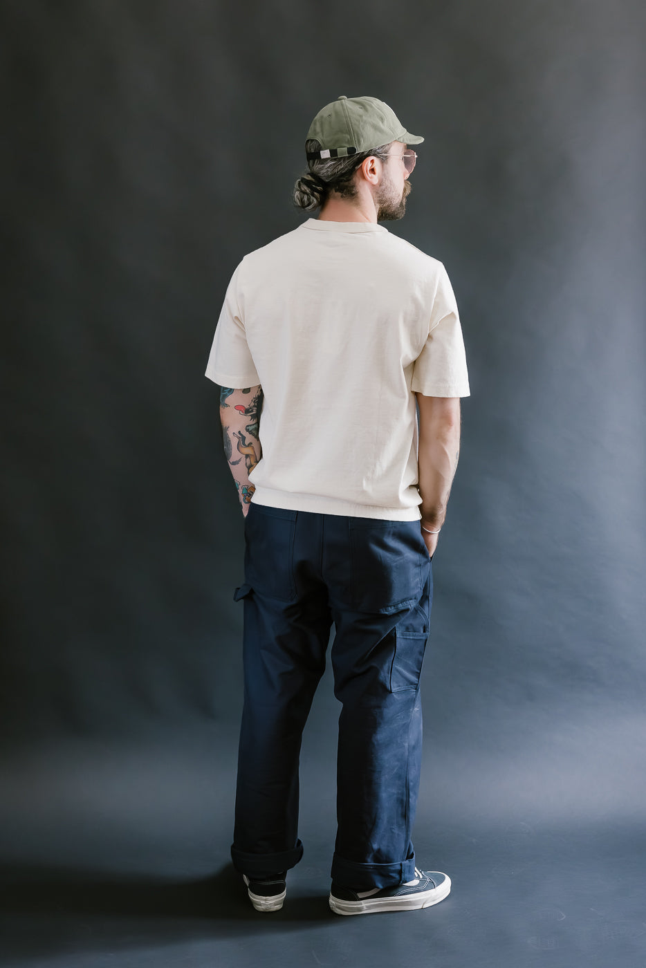 Person wearing a beige t-shirt, navy pants, and a green cap against a gray background