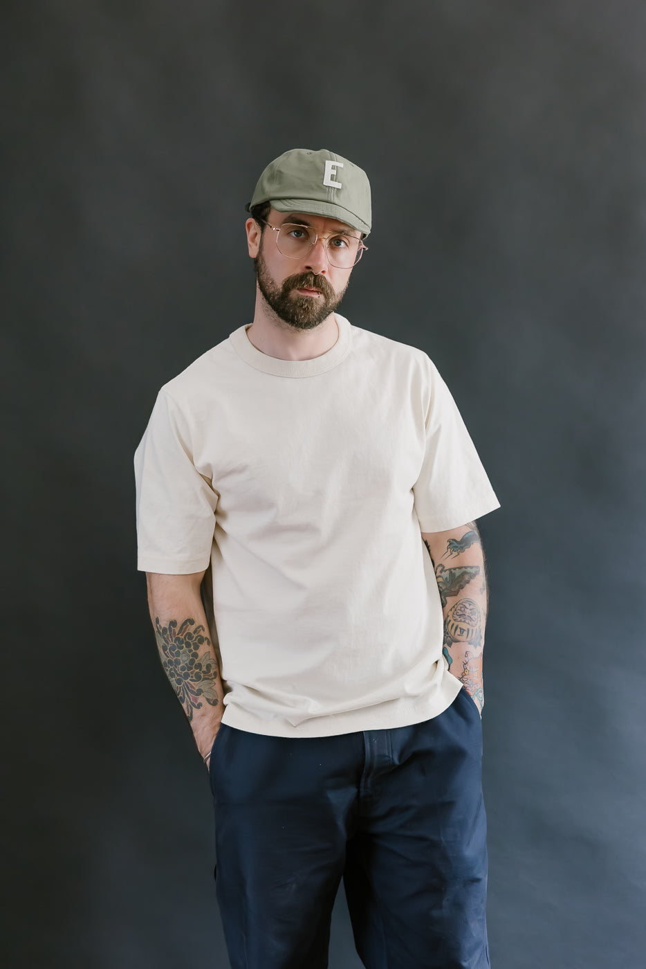 Man wearing a beige t-shirt, navy pants, and a green cap against a dark background