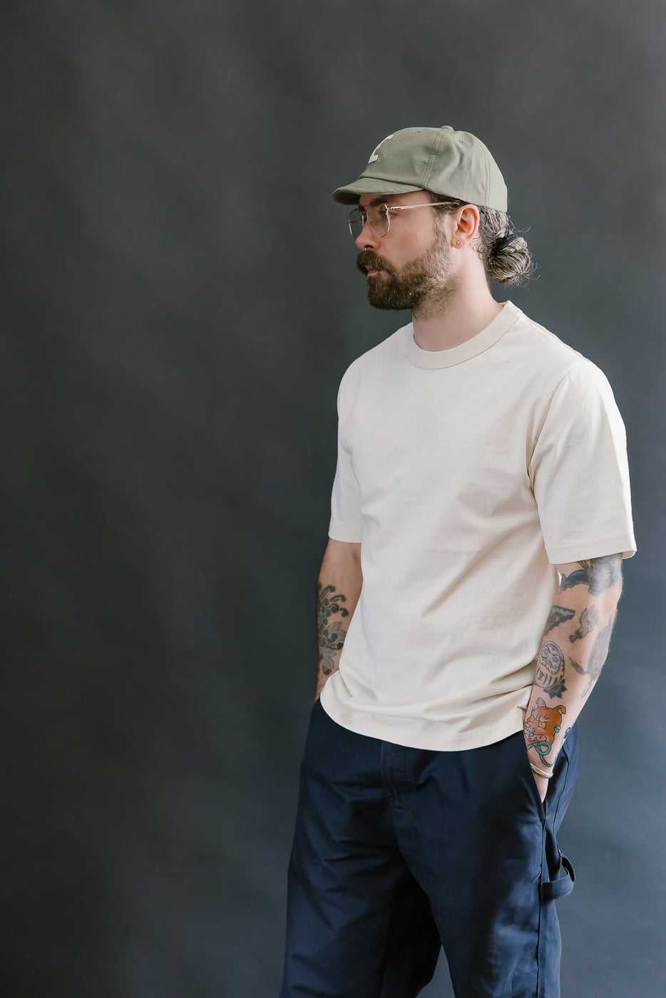 Man wearing a beige t-shirt, navy pants, and a green cap against a dark background