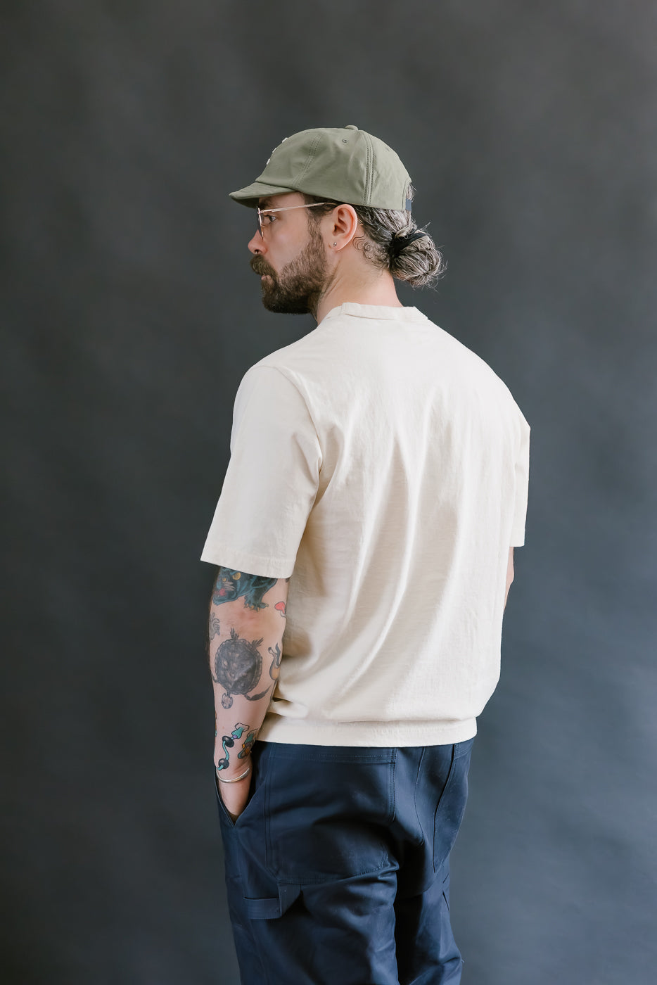 Man wearing a beige t-shirt, navy pants, and a green cap against a dark background