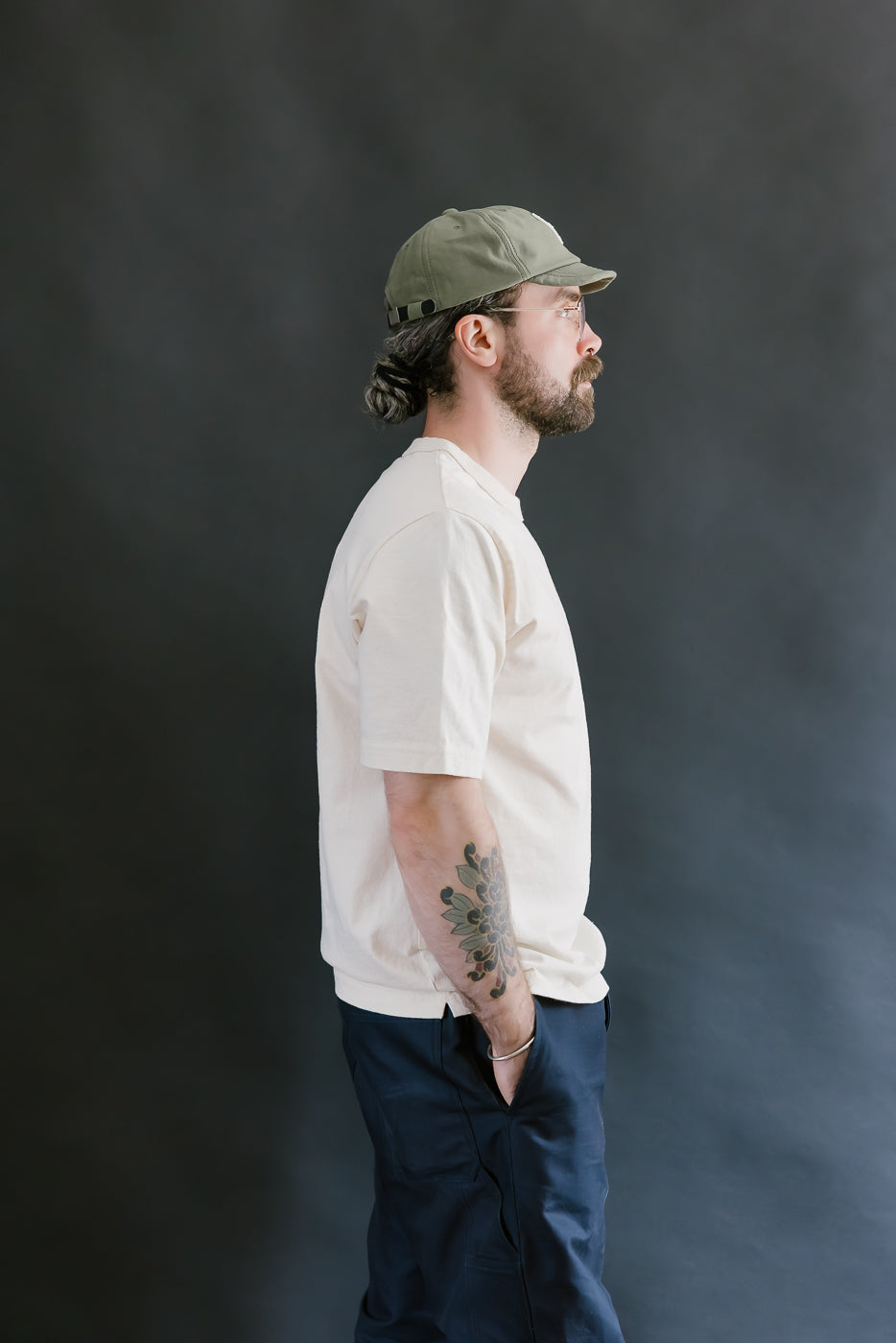 Man wearing a beige t-shirt, dark pants, and a green cap against a dark background