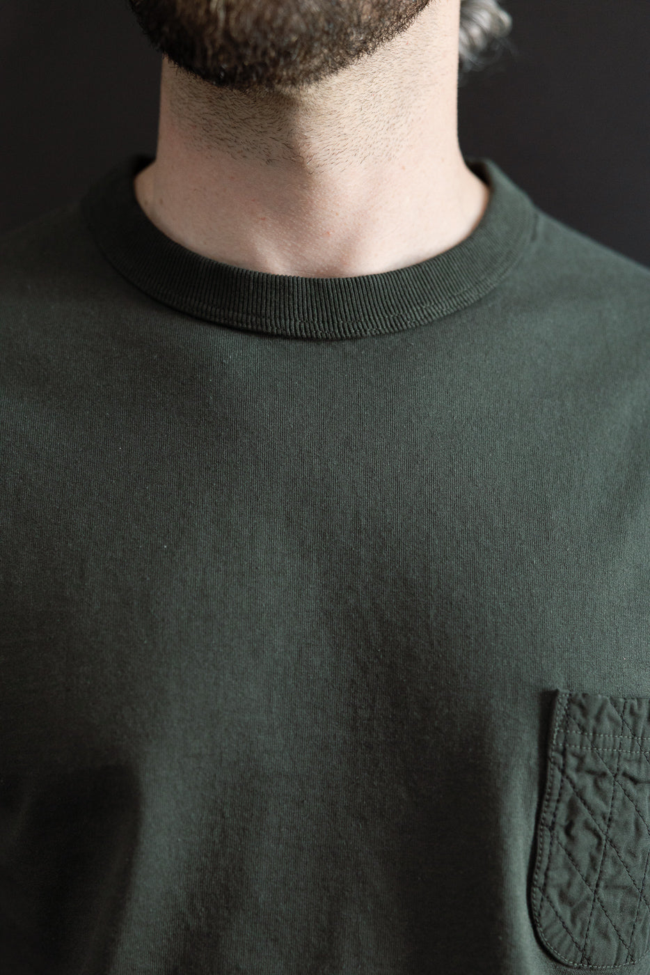 Close-up front view of male model wearing dark green crewneck tee with quilted chest pocket, ribbed collar