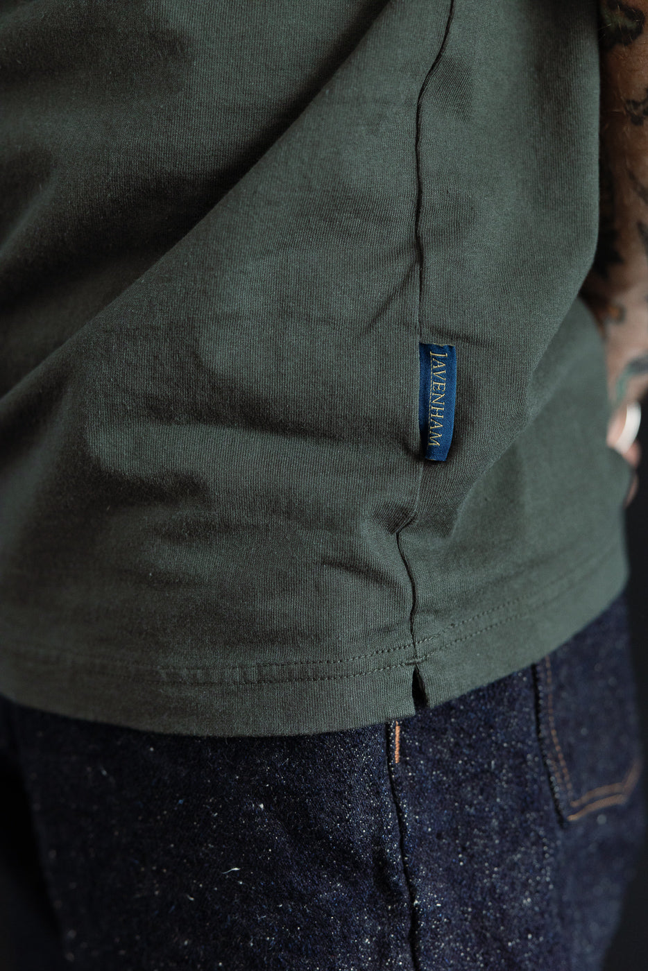 Close-up of olive green tee's side seam and blue Lavenham tag, worn by male model with athletic build