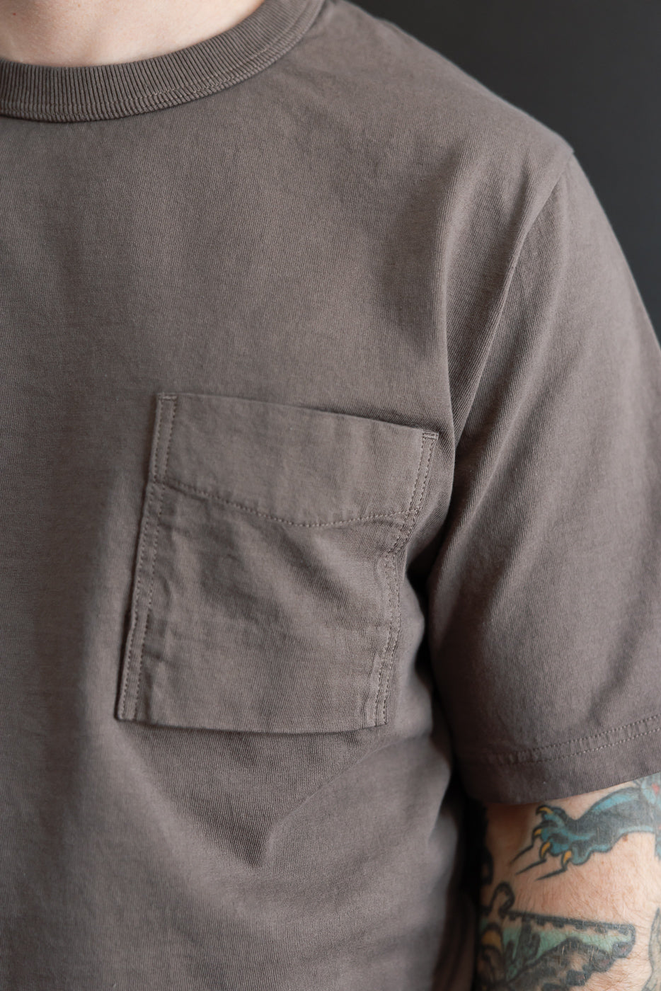 Close-up front view of male model wearing charcoal gray pocket tee, showing chest pocket, ribbed crew neck, and sleeve hem detail.