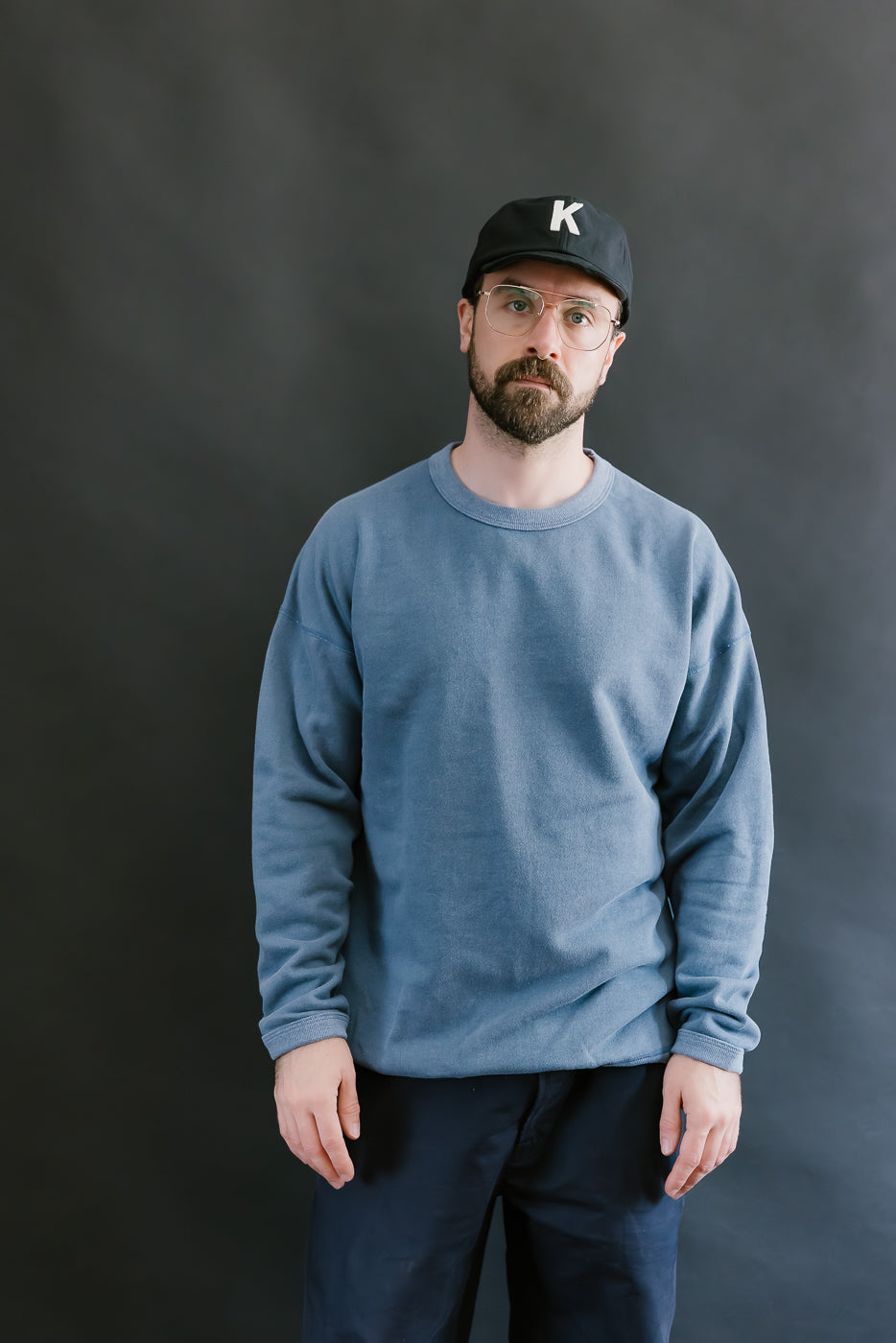 Man wearing a blue sweatshirt and black cap with a 'K' logo against a dark background