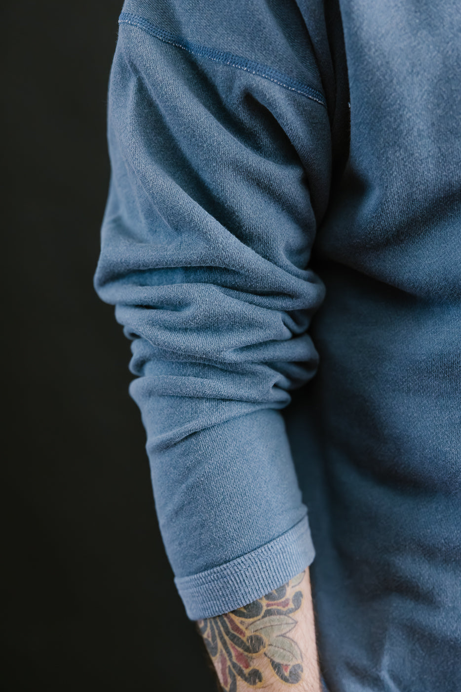 Close-up of a person wearing a blue sweater with a tattoo on the arm against a black background