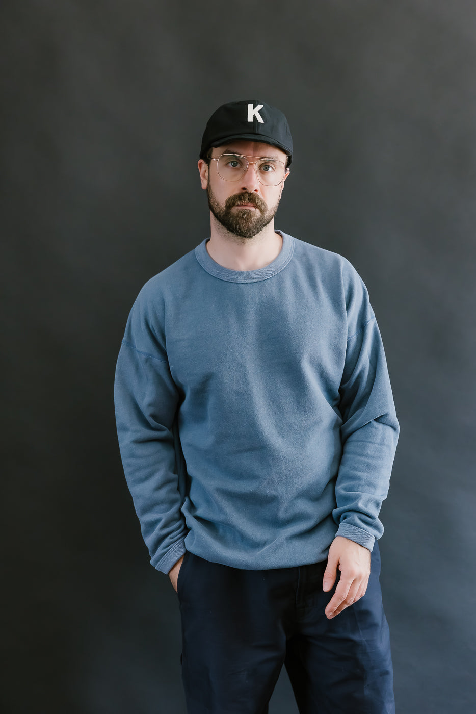 Man wearing a blue sweatshirt and black cap with a 'K' logo against a dark background