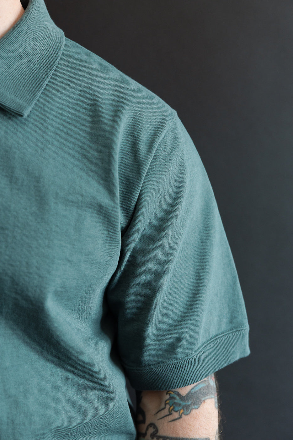 Close-up of male model's shoulder, showing teal polo shirt with ribbed collar and sleeve hem, athletic build