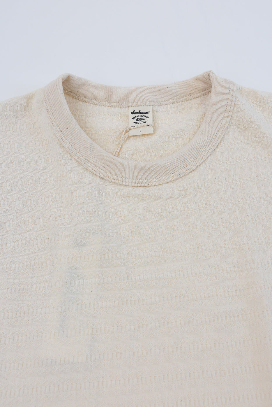 Close-up of front upper chest and collar of ecru textured knit t-shirt, showing tag and ribbed crewneck.