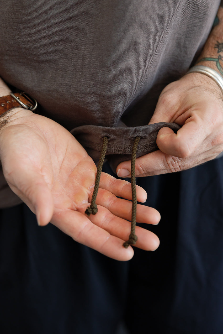 Close-up of male model's hands holding olive drawstring at waist of dark brown hoodie, showing ribbed hem detail