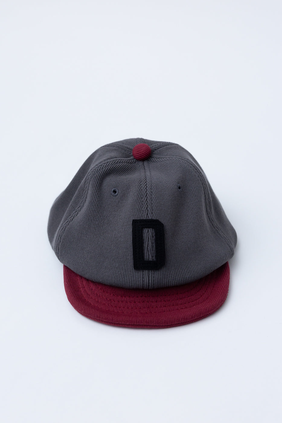 JM6310 - GG Rib BB Cap D features a charcoal gray crown with a burgundy bill and button, highlighted by a black-stitched D on the front, showcasing Jackman's sustainable craftsmanship.