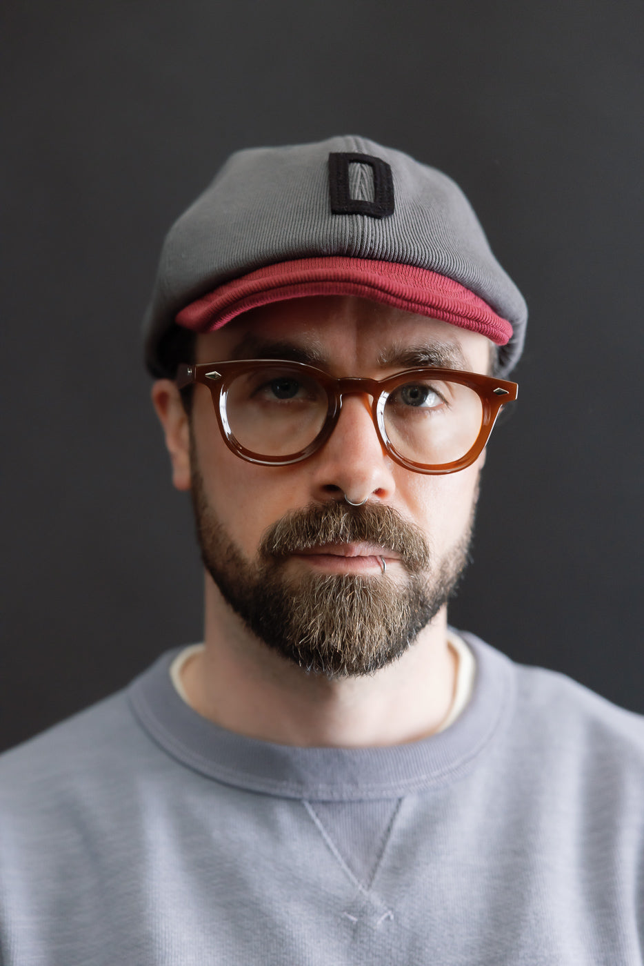 Close-up of a bearded person wearing the JM6310 - GG Rib BB Cap D in 318 Gray x Oxblood, showcasing the cap's ribbed texture and red under-visor, paired with tortoiseshell eyeglasses.